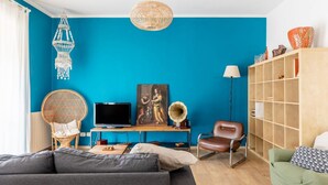Un salon avec un mur bleu, un canapé, un téléviseur, une lampe, une bibliothèque et une suspension décorative.