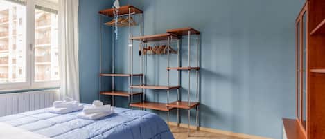 A bedroom with a bed, a wooden shelf unit, a window, and a view of buildings outside.