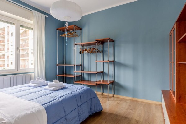 A bedroom with a bed, a wooden shelf unit, a window, and a view of buildings outside.