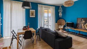 Un salon avec un canapé noir, une table à manger avec des chaises, un téléviseur et une fenêtre avec des rideaux.