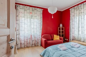 A bedroom with a red wall, a blue bed, a red sofa, a white hanging light fixture, and a wooden nightstand.