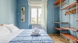 Une chambre à coucher avec un lit, une fenêtre avec des rideaux, un tableau décoratif au mur et une étagère en bois avec des cintres.