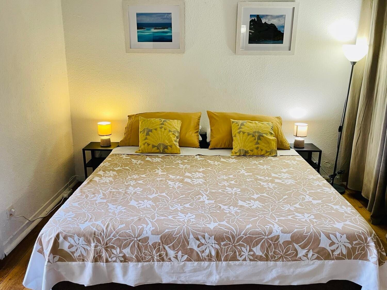 Un lit bien fait, recouvert d’une couvre-lit à motifs floraux, avec deux lampes de chevet et deux tableaux encadrés accrochés au mur.