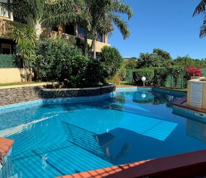 A clear blue swimming pool surrounded by lush greenery and a stone wall. - Studio Hina - Cosy, Quiet & Pool (Papeete)