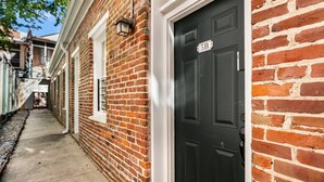 Une ruelle étroite bordée d’immeubles en briques, une porte noire avec une plaque indiquant « 5B », et un rebord de fenêtre blanc.