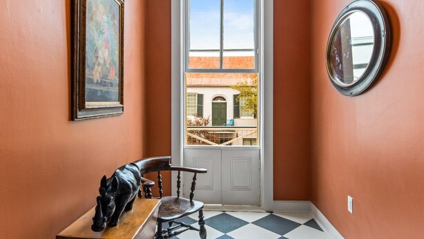 Un corridoio con pavimento a scacchi, una sedia di legno, un quadro incorniciato, uno specchio rotondo e vista su un edificio attraverso una porta.