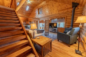 A log cabin interior with a wooden staircase, a sofa, a coffee table, a television, and a fireplace. - Cedars on South: Fall Lakefront Escape | South Manistique Lake | 4BR (Curtis)