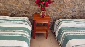 Una habitación con dos camas, una mesita de noche de madera con un florero y una pared de piedra.