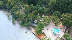 Vue aérienne d’une zone côtière avec une plage, plusieurs petites cabanes et un grand parasol bleu.