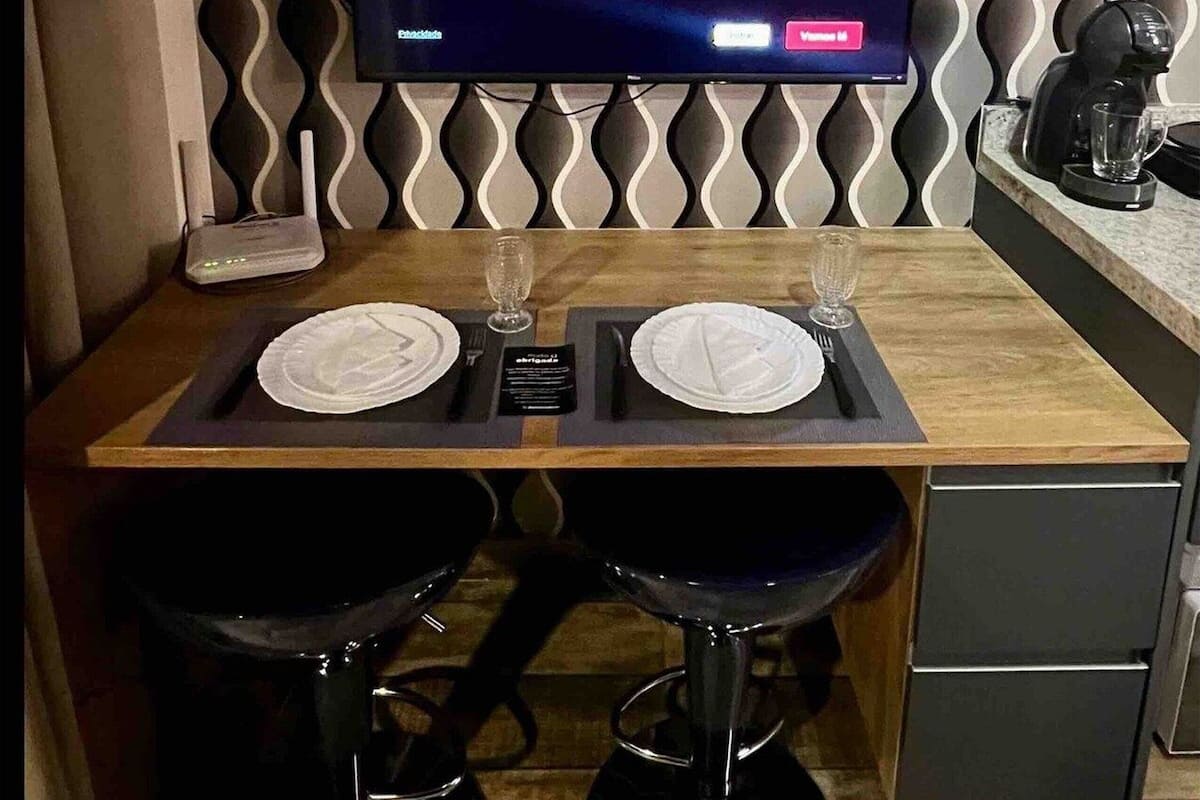 A modern room with a flat-screen TV displaying the Netflix logo, a wooden dining table with two chairs, and a kitchen area with a coffee machine.