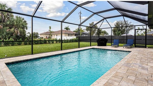 An outdoor swimming pool with a covered area, surrounded by a fence, and a barbecue grill in the background.