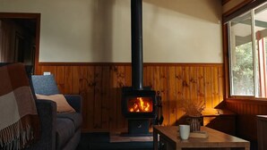 A cozy living room with a wood-burning stove, wooden paneling, a sofa, and a window with a view of trees.