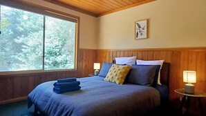A bedroom with a wooden ceiling, a large window, a bed with blue bedding, and a bedside lamp.