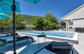 Zona de piscina con sillones de descanso y sombrillas, una mesa pequeña con una copa de vino, y una vista de la montaña al fondo.
