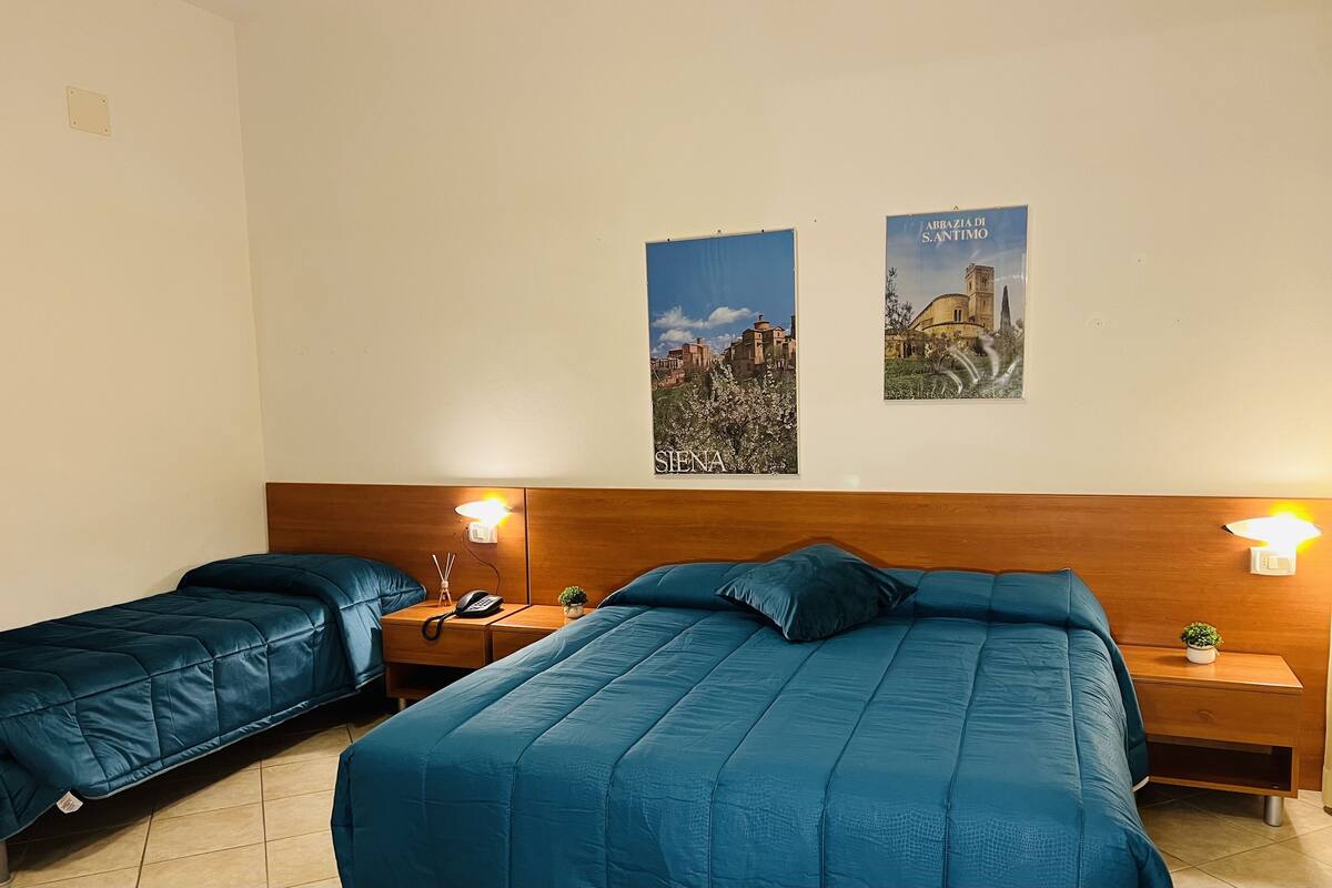 A hotel room with two beds, a wooden headboard, bedside lamps, a small plant, and two posters on the wall.