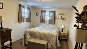 A bedroom with a large bed, a nightstand, a lamp, a potted plant, and a window with curtains. - Spacious 2 bedroom apartment  (Warren)