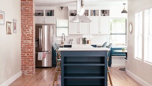 Una cocina moderna con armarios blancos, un pilar de ladrillo, un refrigerador de acero inoxidable y una isla central con encimera oscura.