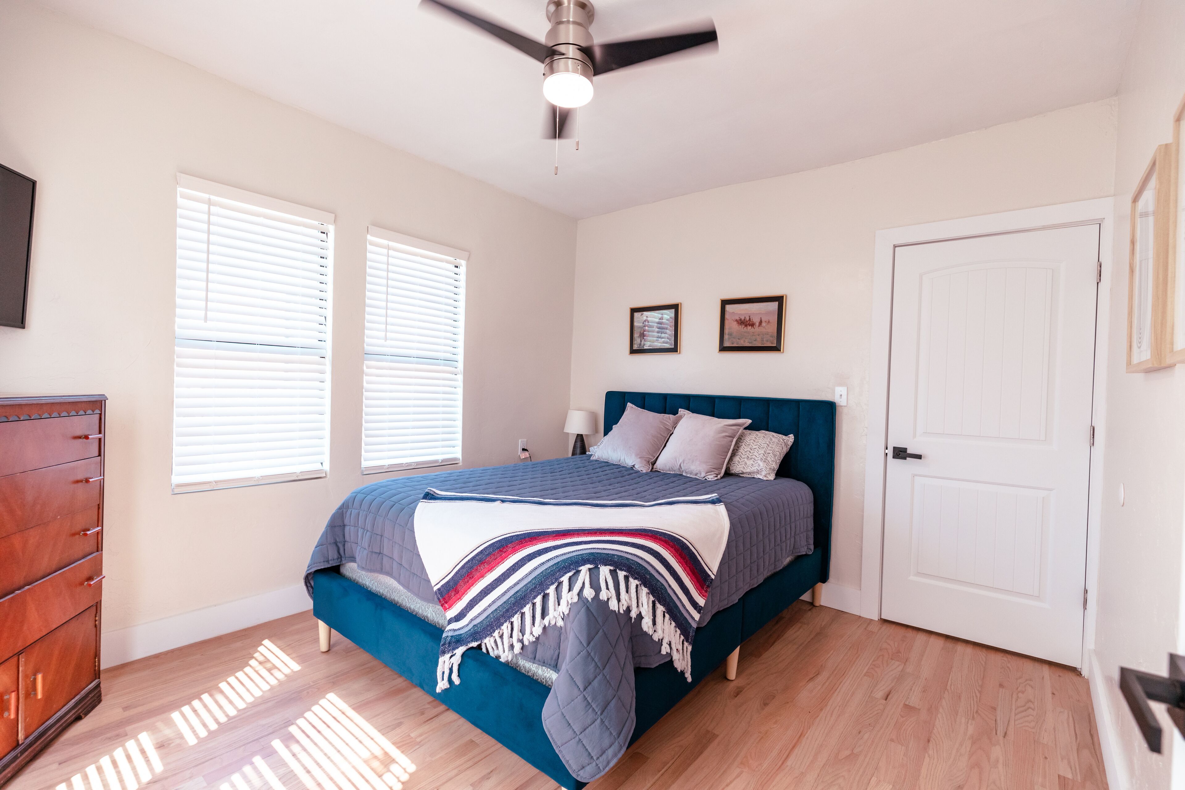 Un dormitorio con cama, cómoda, ventilador de techo y dos ventanas.