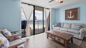 A living room with a sofa, a wooden bench, and a ceiling fan. There is a large window with curtains and a view of the beach.