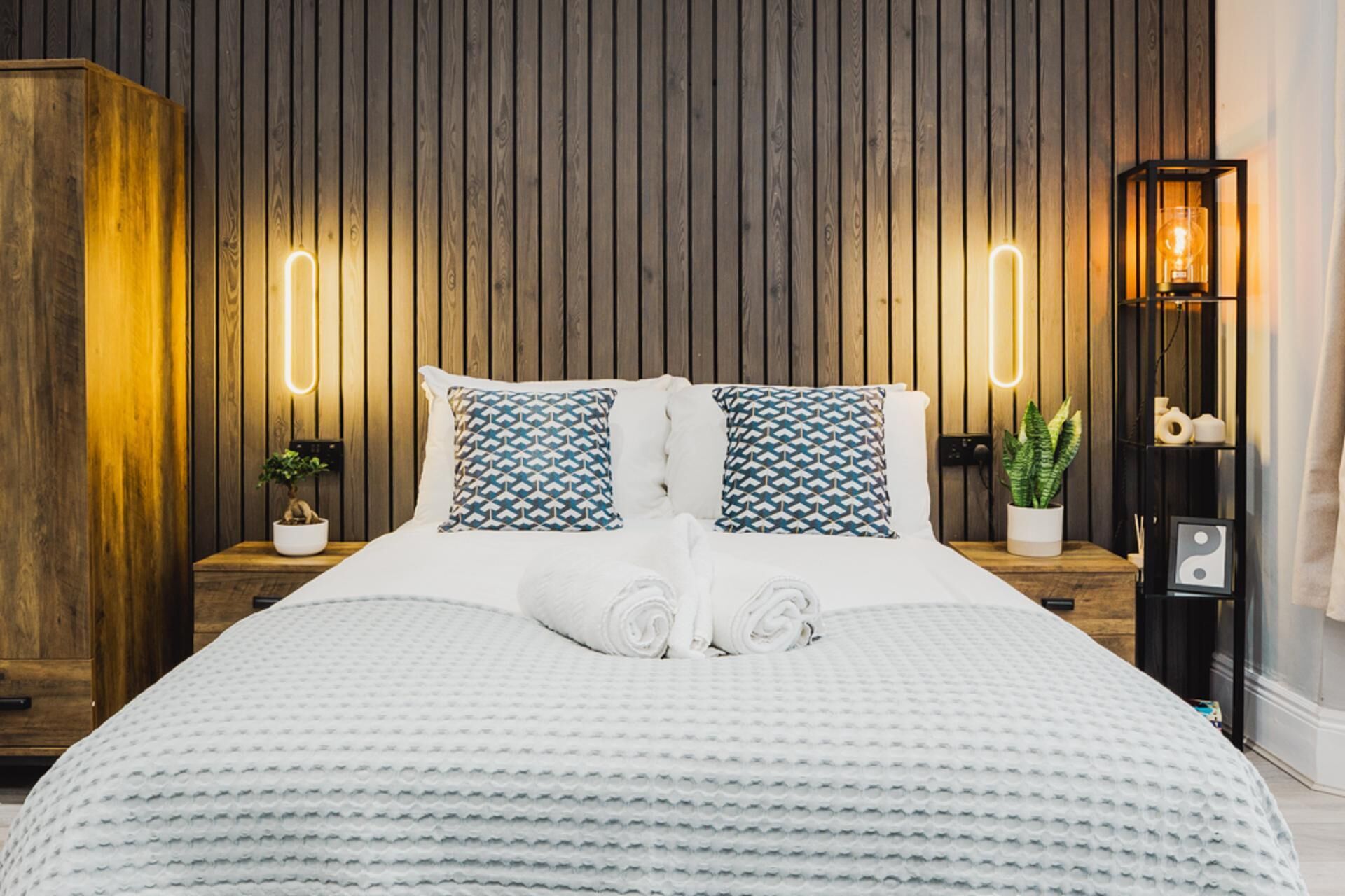 Een netjes opgemaakt bed met een witte deken en twee bedkussens met een patroon, een kamerplantje en een wandlamp.