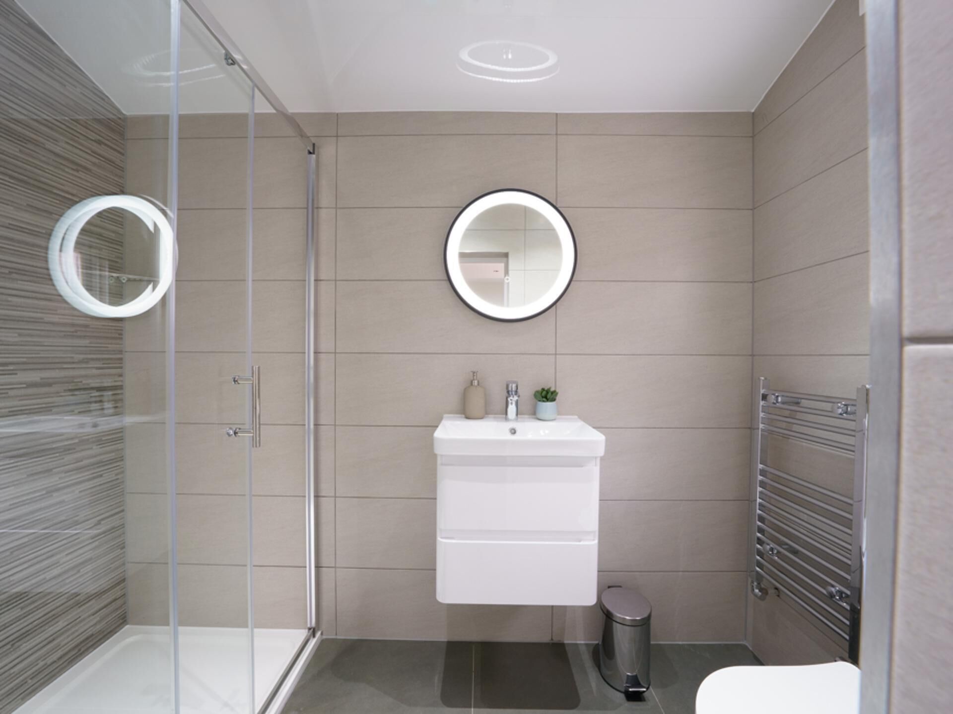Un bany modern amb una cabina de dutxa de vidre, un lavabo blanc amb un mirall a la part superior i un escorredor de tovalloles a la dreta.