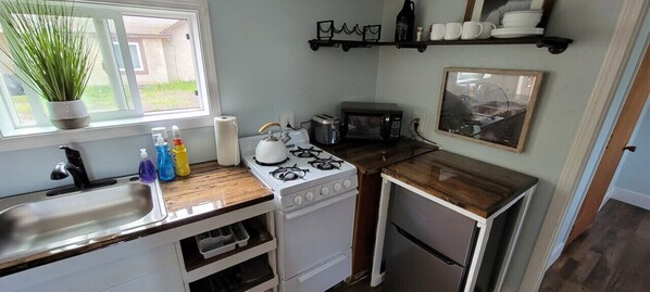 A compact kitchen with a gas stove, microwave, and a small refrigerator. There is a sink with dish soap and a potted plant by the window.