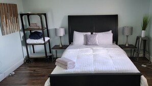 A neatly made bed with a white comforter and pillows, two bedside lamps, a small side table with a lamp, and a wooden shelf unit with folded towels. - 10 minute walk to restaurants & trails - Convenient Location - #4 Slumber Village (Anchorage)