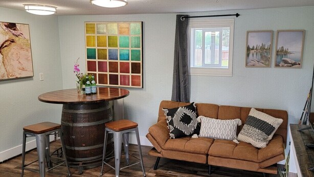 A living room with a brown sofa, a round wooden table, and bar stools. There are paintings on the walls and a window with a view. - 10 minute walk to restaurants & trails - Convenient Location - #4 Slumber Village (Anchorage)
