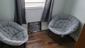 A corner room with two grey cushioned chairs, a framed picture on the floor, and a window with curtains. - 10 minute walk to restaurants & trails - Convenient Location - #4 Slumber Village (Anchorage)