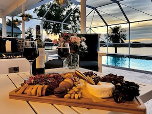 Uma mesa com tábua de frios, garrafa de vinho, copos e flores junto a uma piscina.
