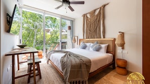 A bedroom with a bed, a desk, a chair, a ceiling fan, a TV, and a large window with a view of trees. - NHOA 207B Lux 1BR Condo Pool View in Aldea Zama (Tulum)