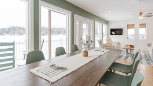 A dining area with a long wooden table, green chairs, and a view of a snowy landscape through large windows. - Lakefront Family Fun Retreat (Newport)