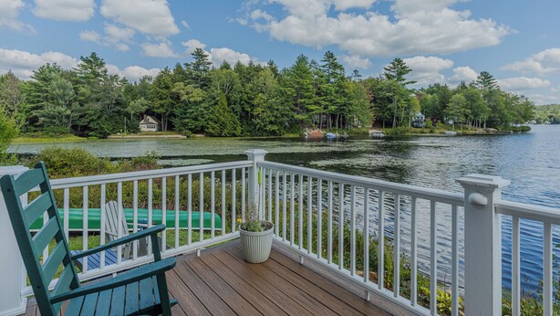 A wooden deck with a white railing overlooking a lake, surrounded by trees and a small house. - Lakefront Family Fun Retreat (Newport)