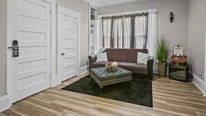 Una sala de estar con un reposapiés verde oscuro, un sofá marrón y una ventana con cortinas.