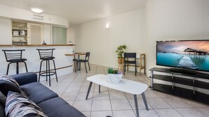 Una sala moderna con un sofá, un comedor con sillas, una cocina con mostrador y armarios, y un televisor que muestra una vista panorámica.