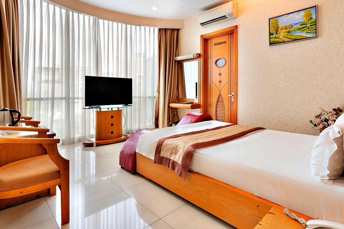 A hotel room with a bed, a desk with a chair, a television, and a window with curtains.
