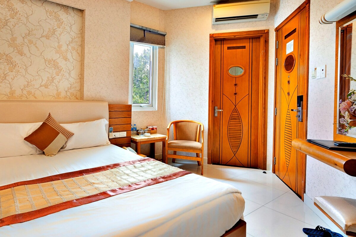 A hotel room with a large bed, wooden headboard, a small table with a chair, a wooden wardrobe, and a window with a view of trees.