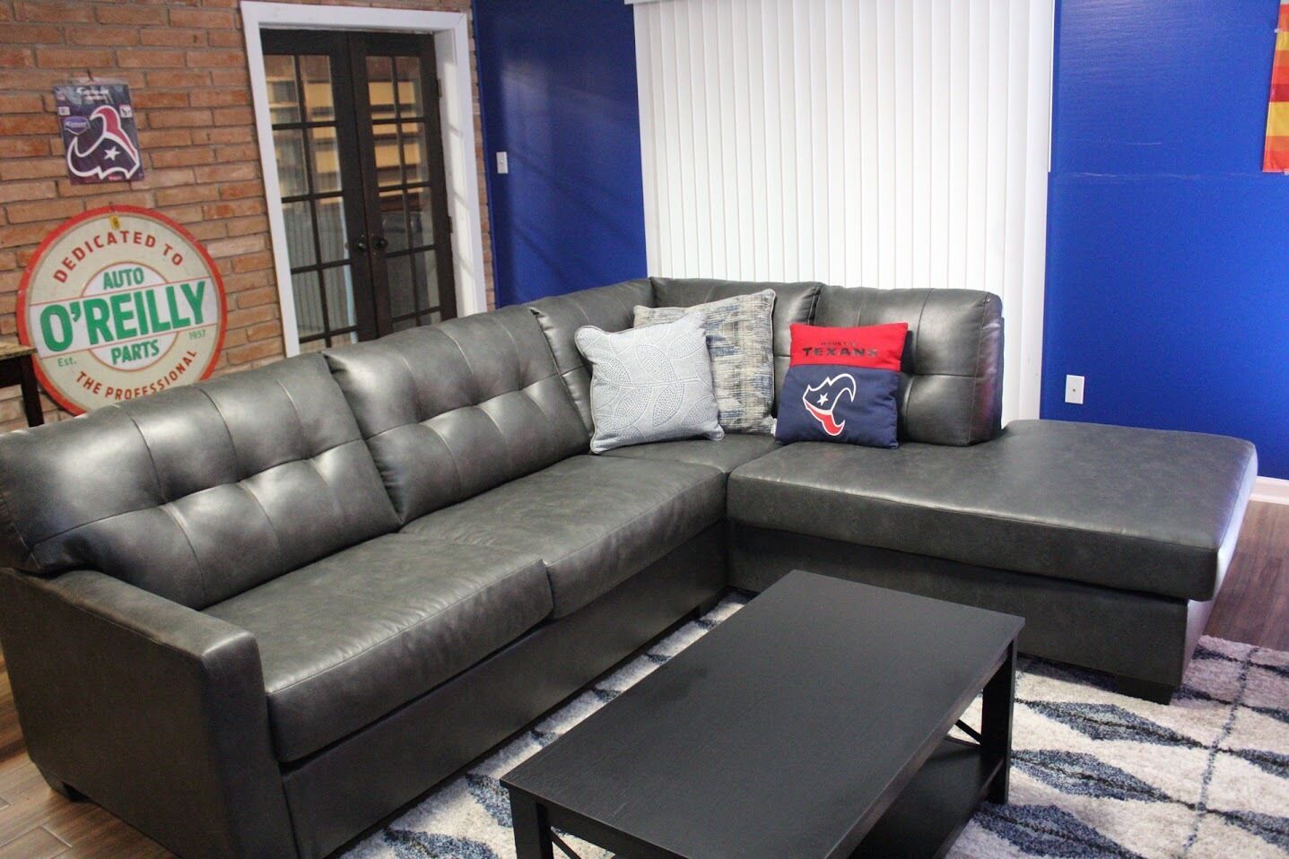 Un canapé d’angle en cuir noir, accompagné d’un pouf assorti et d’une petite table basse, dans une pièce aux murs bleus et dotée d’un mur d’accent en briques.