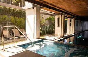 Piscina interior moderna con paredes de vidrio, techo de madera y vistas al exterior con vegetación.
