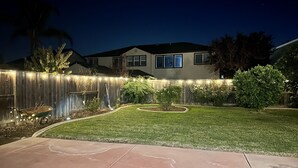 A residential backyard at night with a well-lit fence, a lawn, and a house in the background. - Discover Turlock charm near California State University  (Turlock)