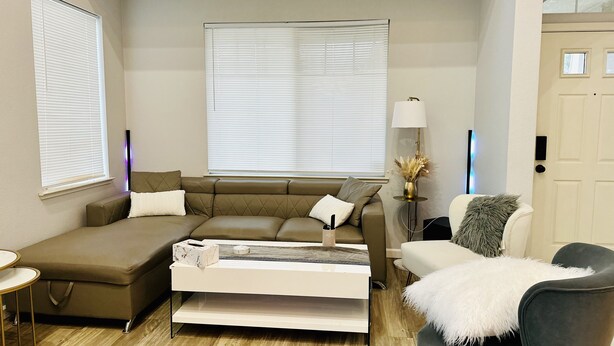 A modern living room with a sectional sofa, a coffee table, and a side table. There are two windows with blinds, a lamp, and a door. - Discover Turlock charm near California State University  (Turlock)