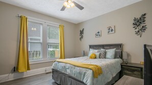 A bedroom with a bed, a nightstand, a lamp, a ceiling fan, and a window with yellow curtains. - Your Newest, Remodeled, and Fully-Furnished AirB&B in the Heart of Fayetteville! (Fayetteville)