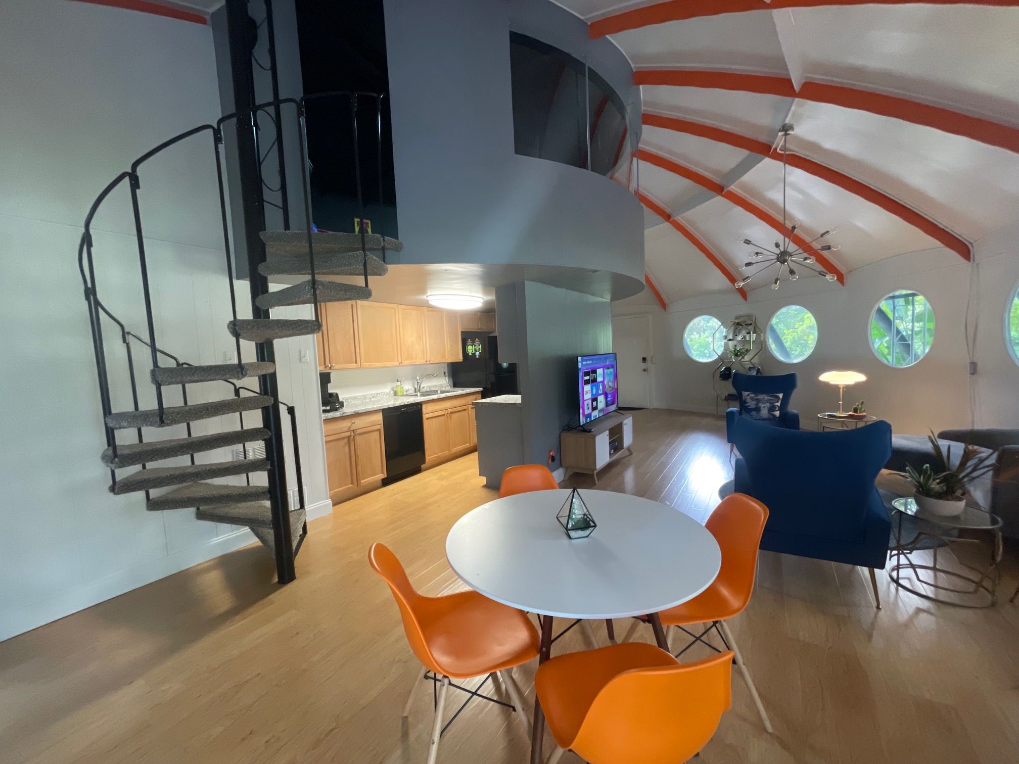 A modern interior with a spiral staircase, a dining area with orange chairs, and a kitchen in the background.
