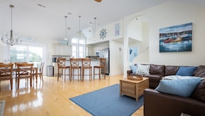 A spacious living room with a brown leather sofa, wooden dining table, and chairs. A kitchen with white cabinets and a dining area with a blue rug is visible in the background. - 3min Walk to Beach, Pool, Hot Tub, & Game Room! (Corolla)