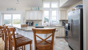 A kitchen with white cabinets, a stainless steel refrigerator, a coffee maker, and a bar area with wooden chairs. - 3min Walk to Beach, Pool, Hot Tub, & Game Room! (Corolla)