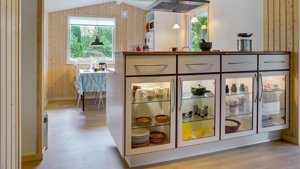 Et moderne køkken med træpaneler, en central ø med skabe med glaslåger, og et spiseområde med et bord og stole.