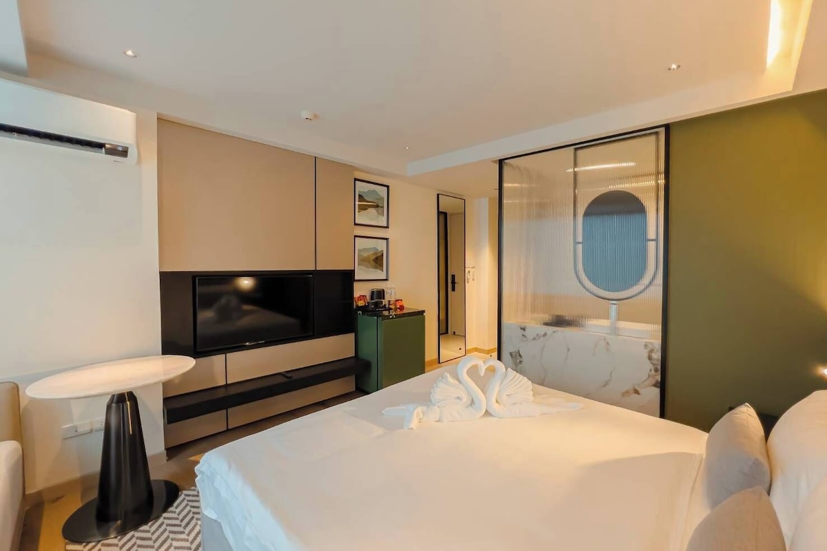Quarto de hotel moderno com uma cama grande, uma TV de tela plana, uma mesinha e um espelho.