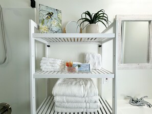 Un baño con un estante blanco que contiene toallas, una planta en maceta y una obra de arte enmarcada.