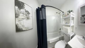 Een badkamer met douche, toilet en wastafel. Er hangt een schilderij aan de muur en een plank met handdoeken.