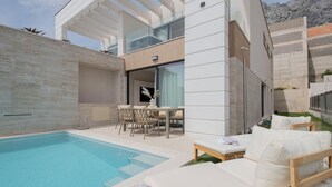 A modern outdoor pool area with a lounge chair and a table, surrounded by a minimalist architectural design.